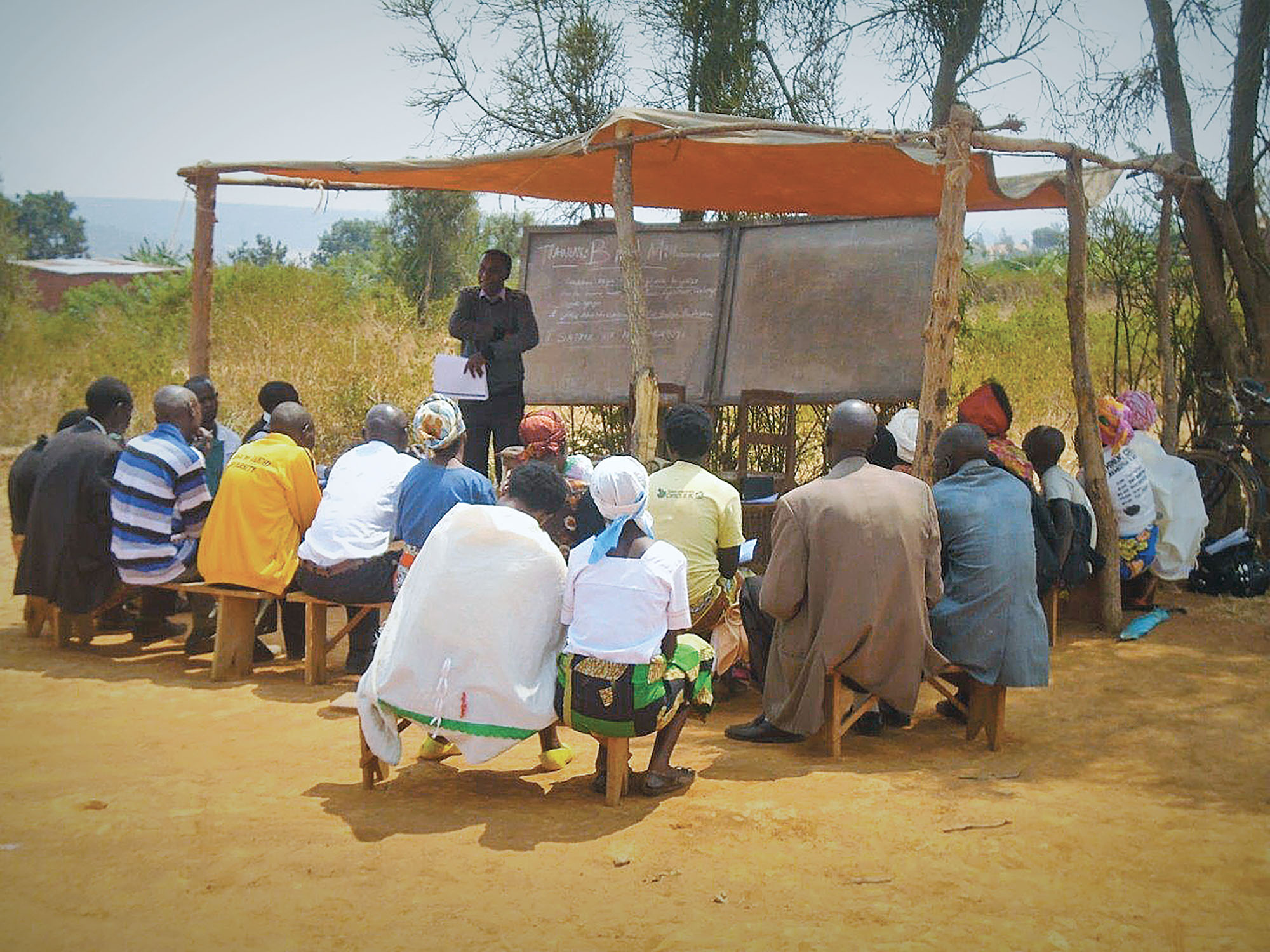 A man teaches a group of adult students in an outdoor classroom in Africa.