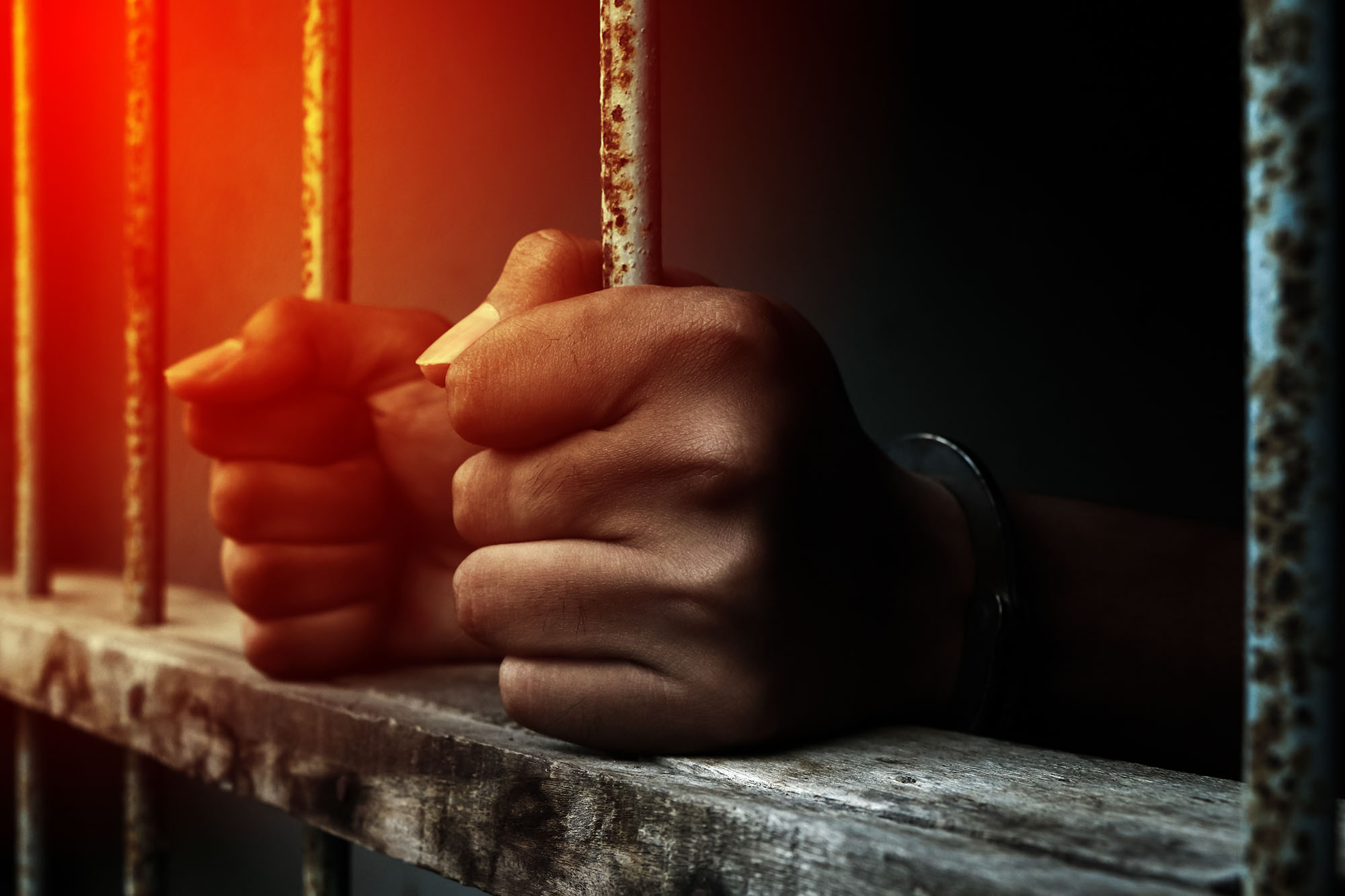 A person's hands grip the bars of a prison cell.